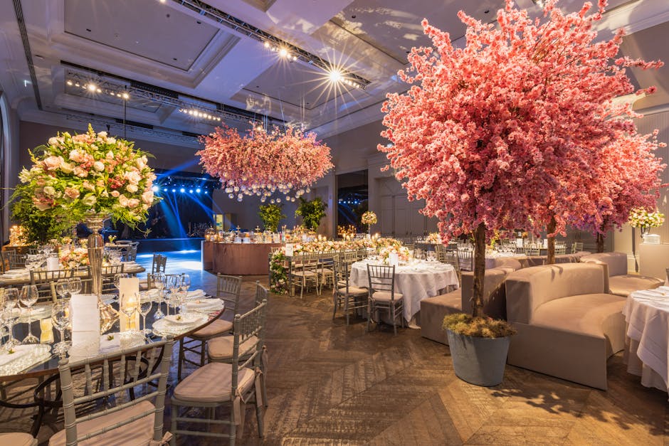 An elegantly decorated event space featuring a large pink flowering tree in a gray pot positioned beside a plush beige sofa. The room is illuminated by soft lighting, with additional bright spotlights