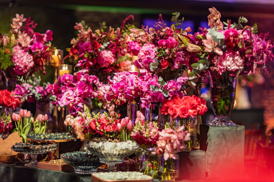 A visually striking floral display featuring a variety of fresh, vibrant pink and red flowers arranged in tall, elegant glass vases and decorative bowls. The arrangement includes orchids, roses, tulip