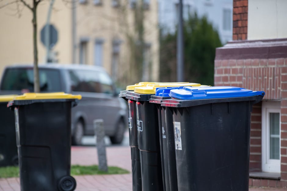 A row of four black waste bins with yellow and blue lids positioned on a sidewalk in front of a brick building. The bins are lined up next to each other, with two sharing blue lids, one with a yellow 