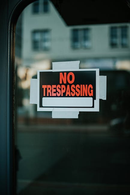 The image features a 'No Trespassing' sign attached to a glass surface with white tape. The sign has a black background with bold red lettering for 'NO TRESPASSING' and a white blank space underneath 