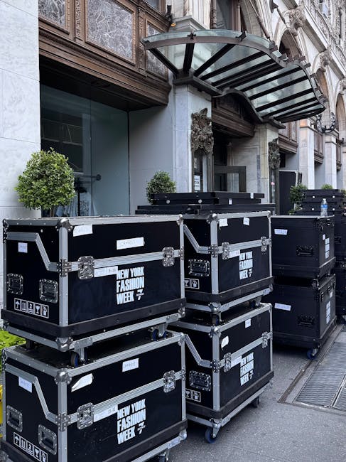 A row of black flight cases labeled 'New York Fashion Week' stacked outside a building with ornate architecture, featuring large glass windows, a modern awning, and small, neatly trimmed green shrubs 