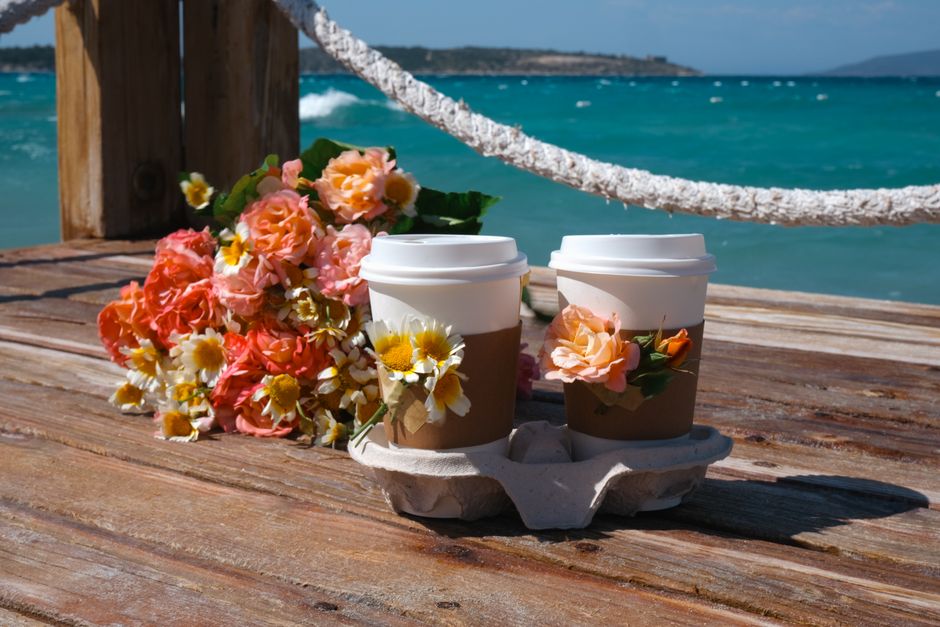 A floral arrangement featuring a bouquet of pink and peach roses, white daisies with yellow centers, and green foliage resting on a weathered wooden surface near the seaside. Beside the bouquet, there