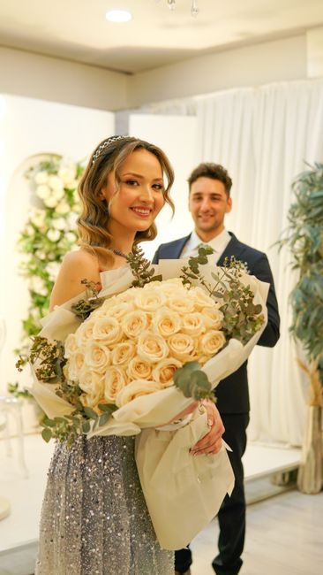 A woman with light brown, shoulder-length hair styled in loose waves and wearing a delicate headband, is holding a large, elegant bouquet of cream-colored roses accented with sprigs of eucalyptus and