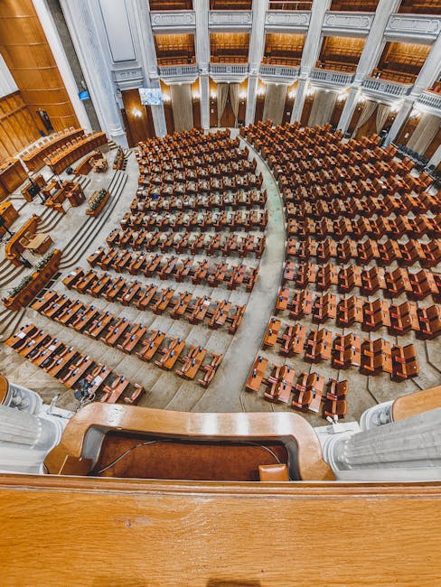 An expansive view of a grand, historic assembly hall with rows of neatly arranged wooden desks and chairs, leading up to a stage with a grey podium and ornate white columns flanking the front. The roo