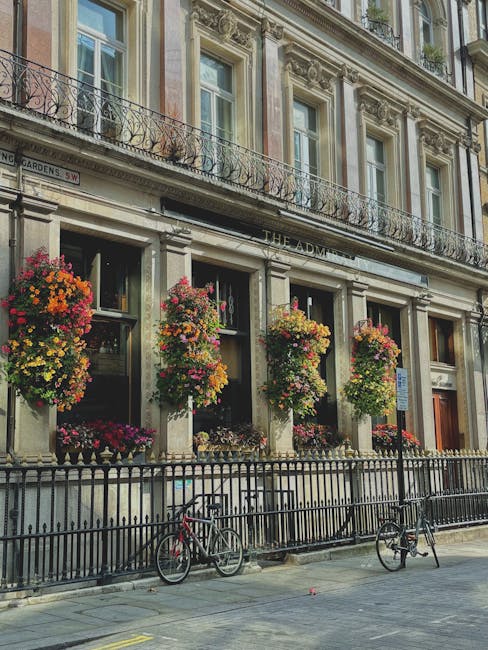 The image showcases the exterior of a classic London building with ornate architectural details and large windows, decorated with vibrant hanging floral baskets filled with multicolored flowers includ